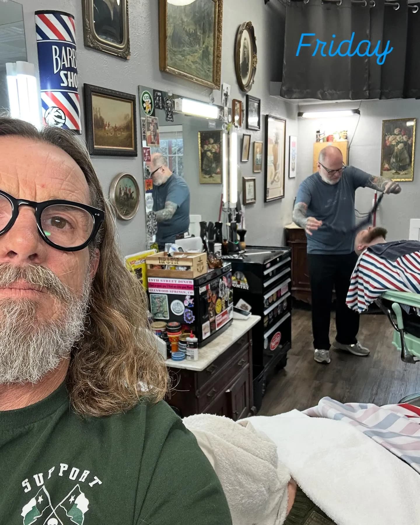 Jake in a selfie at the Wimberley barber shop with a barber cutting a client's hair behind him