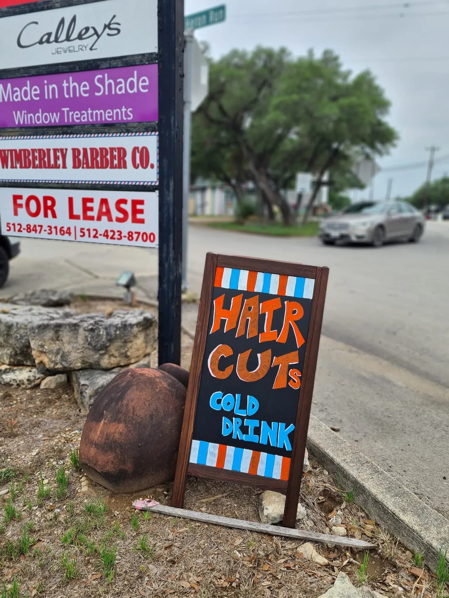 Hand-painted sandwich board reading Hair Cuts and Cold Drink outside Wimberley Barber Co.